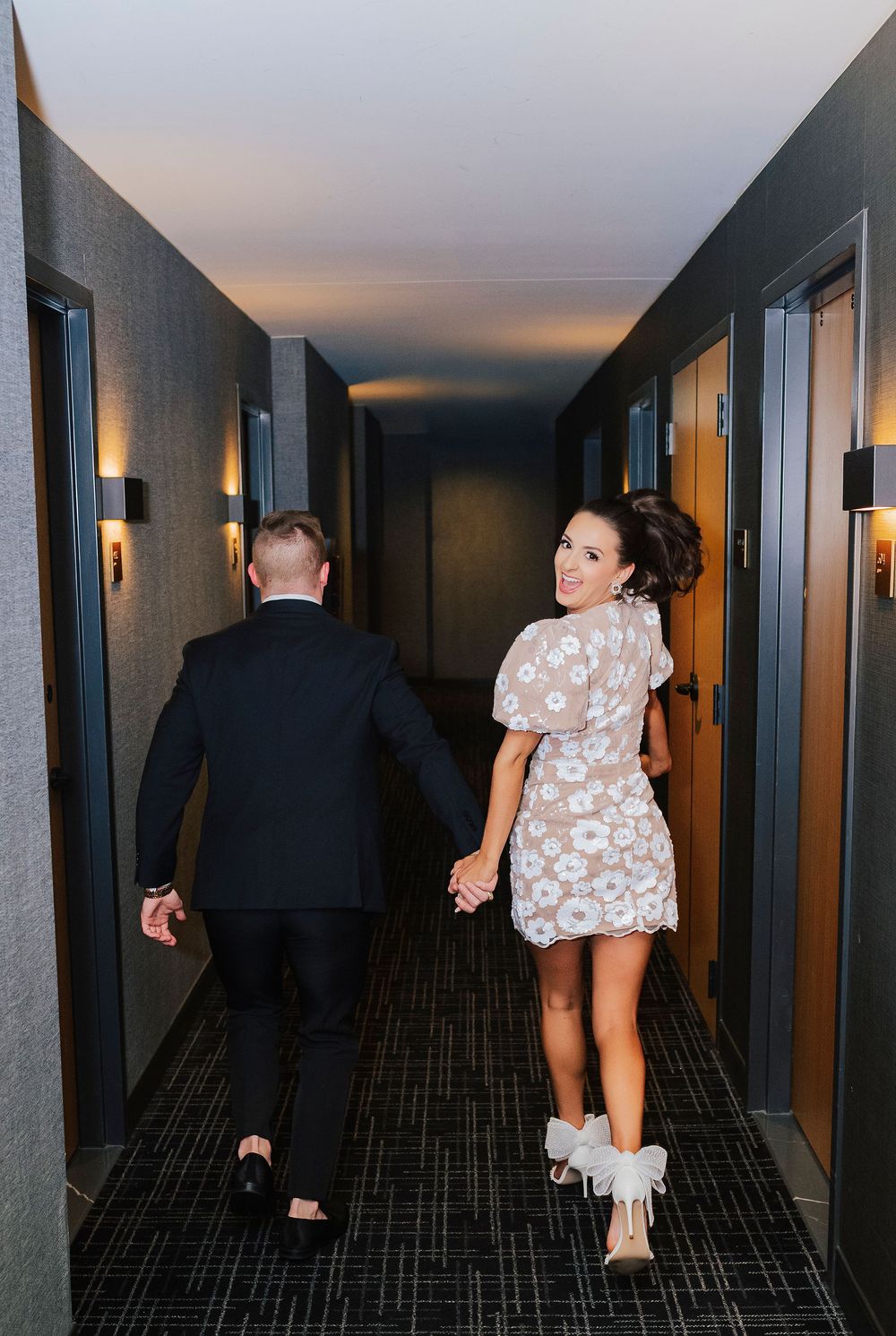 Couple walking hand-in-hand down a dimly lit hotel hallway, woman in floral mini dress glancing back smiling.