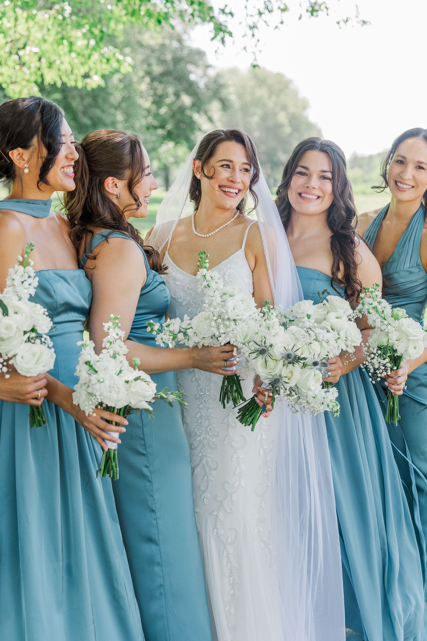 Smiling bride in white lace gown and veil holds bouquet surrounded by bridesmaids in dusty blue dresses outdoors.