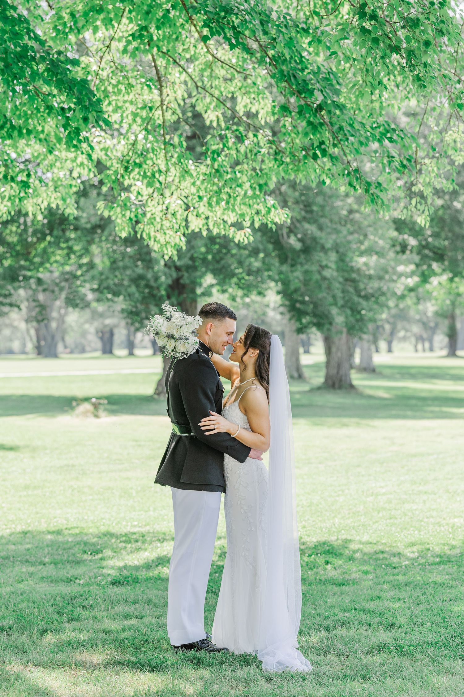 Bride and groom share a romantic moment under lush green trees in a sunlit park, bride holding white flowers.