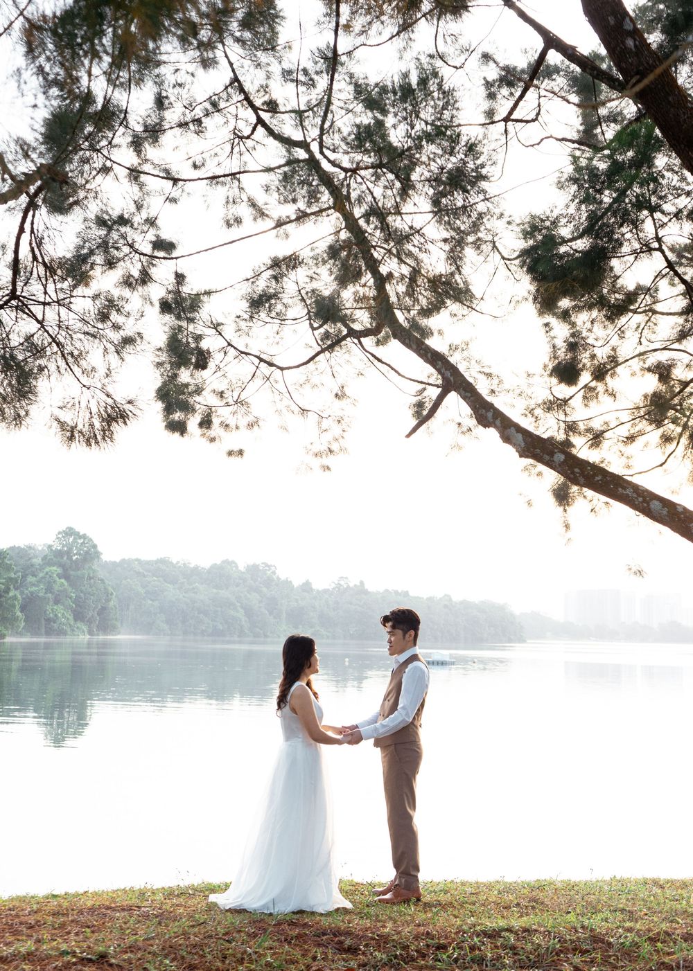 Couple holding hands in wedding attire by a serene lake under pine trees at sunset.
