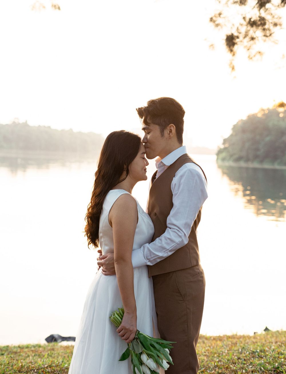 Couple sharing a romantic moment by a serene lake at sunset, bride in white dress holding tulips, groom in brown vest.