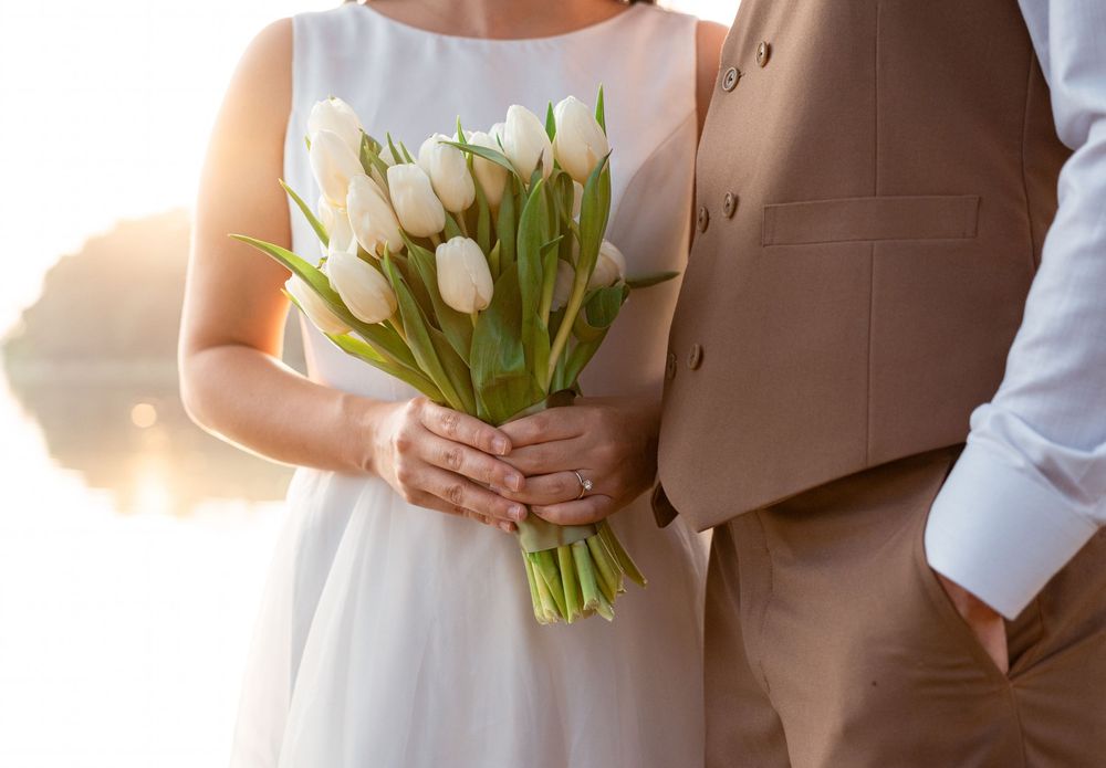 Bride holding white tulip bouquet with groom in tan suit at golden hour wedding ceremony.