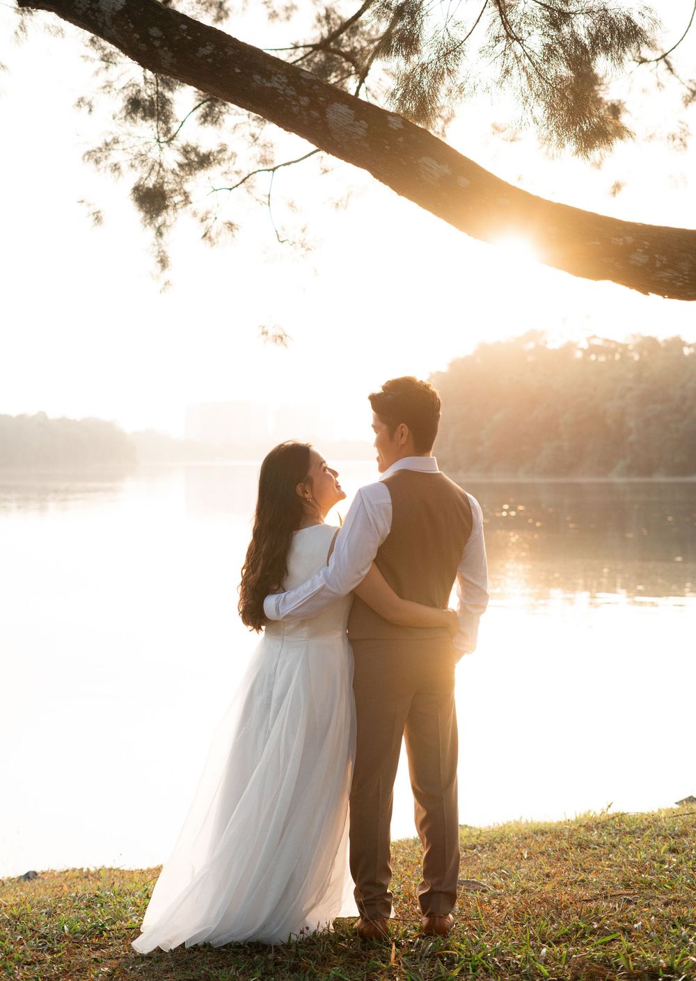 Couple in wedding attire embracing by a sunlit lake under pine trees at golden hour.