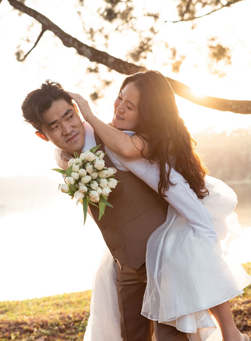 Couple in wedding attire sharing a romantic moment outdoors at golden hour, groom holding white tulip bouquet.
