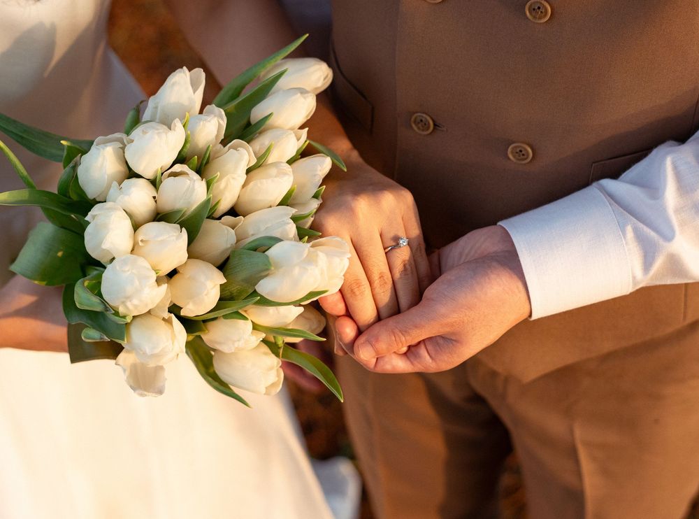 Bride and groom holding hands with white tulip wedding bouquet, featuring a diamond engagement ring.