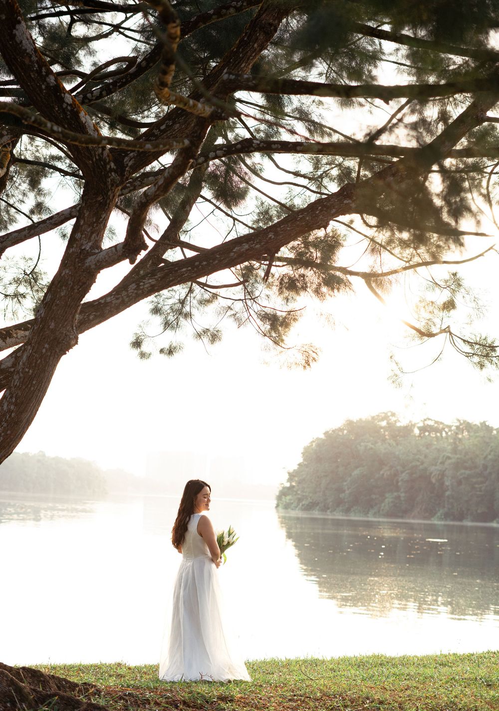Bride in white gown holding bouquet standing by a serene lake under pine trees at sunset.
