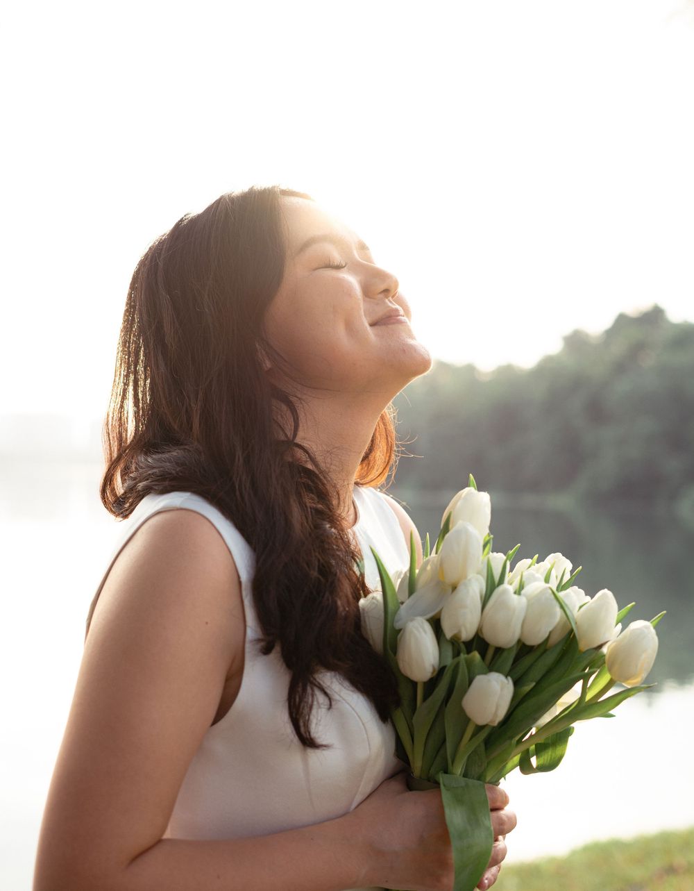 Young woman in white dress joyfully holding bouquet of white tulips outdoors near water in soft sunlight.