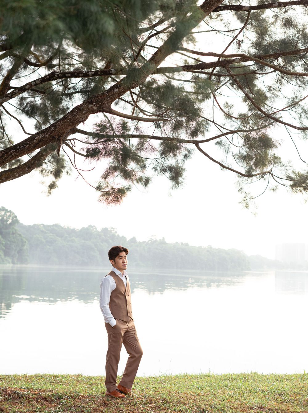 Man in vest and trousers standing by misty lake under pine tree branches in serene outdoor setting.