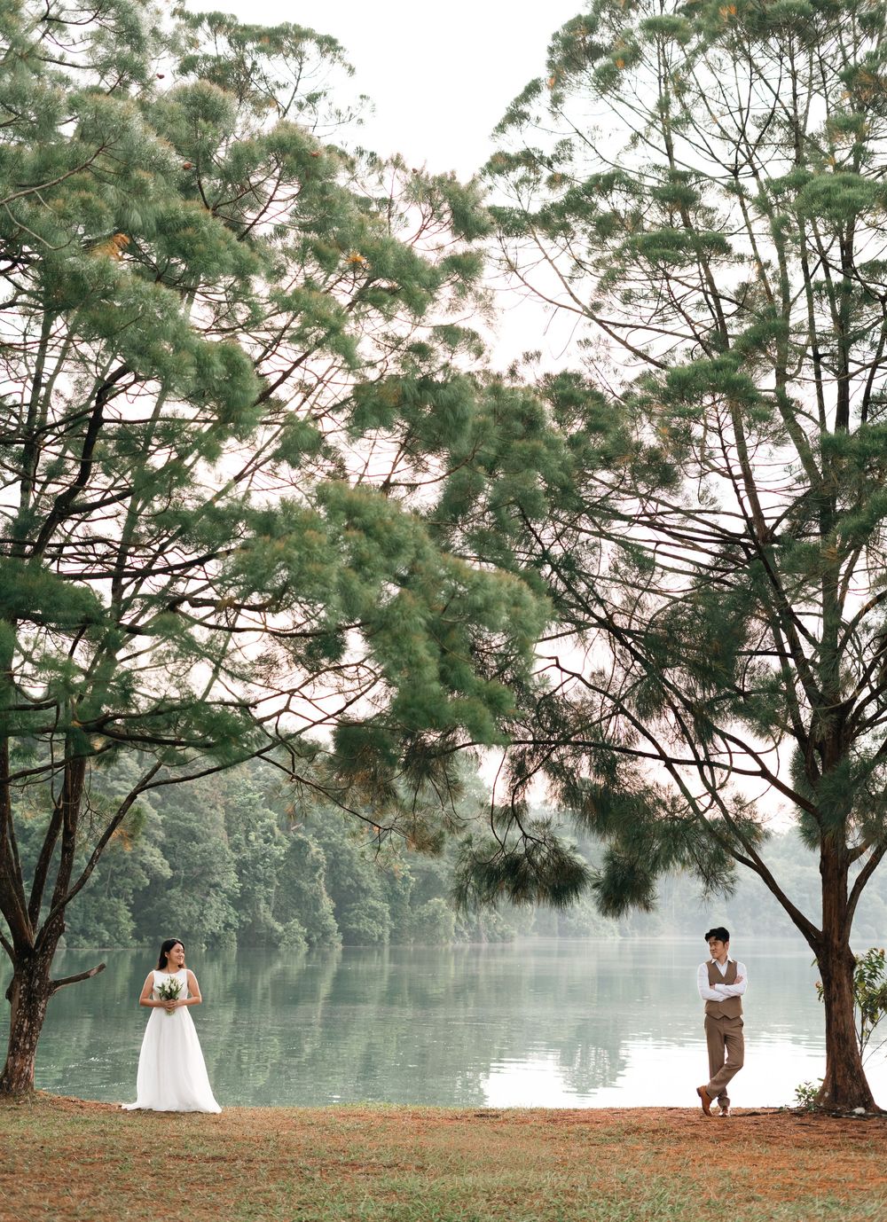 Bride and groom stand apart near a serene lake surrounded by tall pine trees during an outdoor wedding photoshoot.