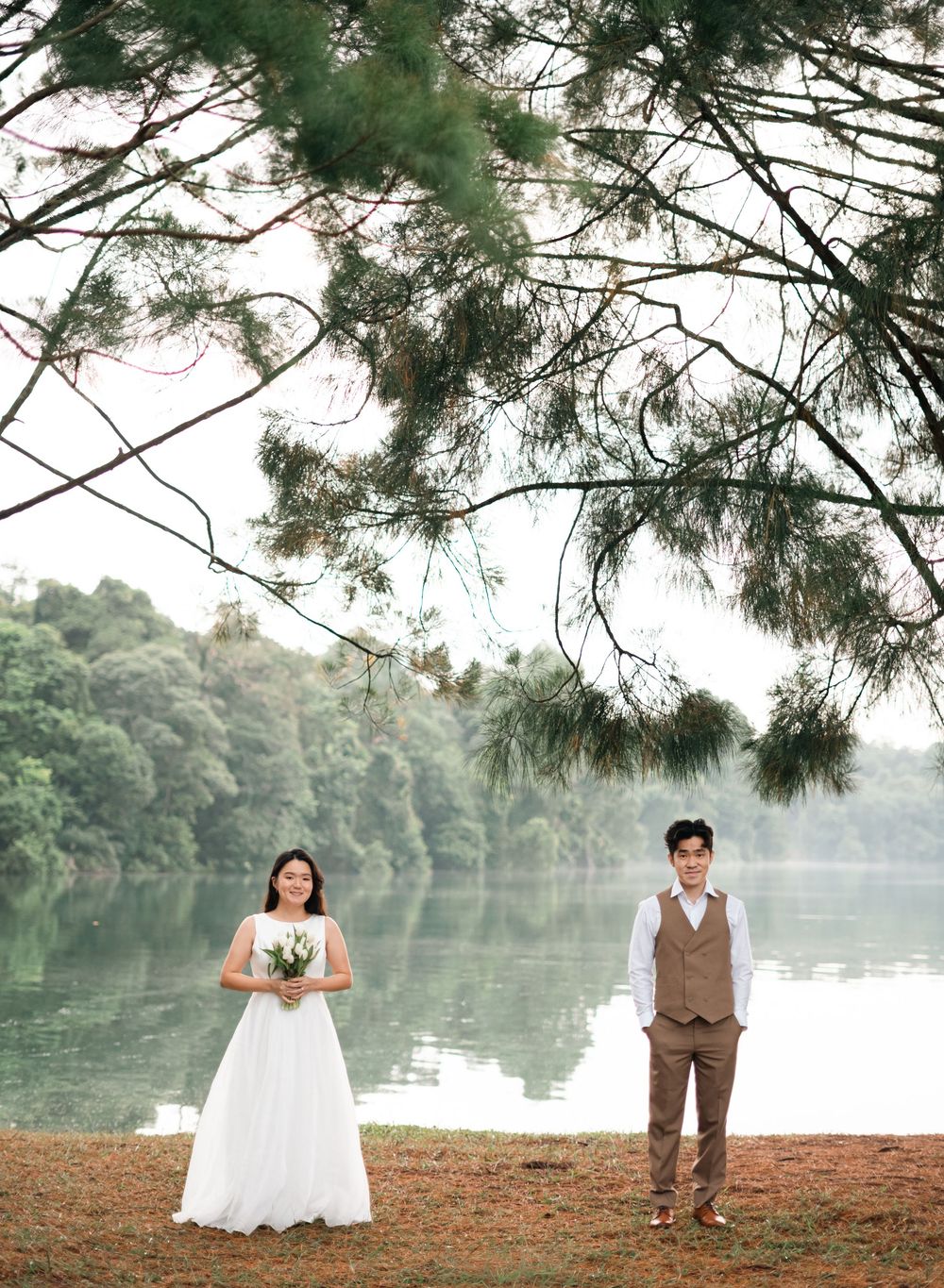 Bride in white gown and groom in brown suit stand apart by a serene lake surrounded by lush greenery and pine trees.