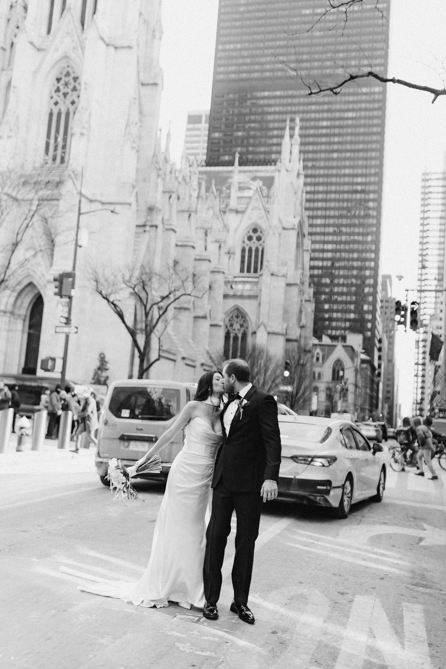 Black and white photo of a bride and groom kissing on a NYC street with St. Patrick's Cathedral and skyscrapers in background.
