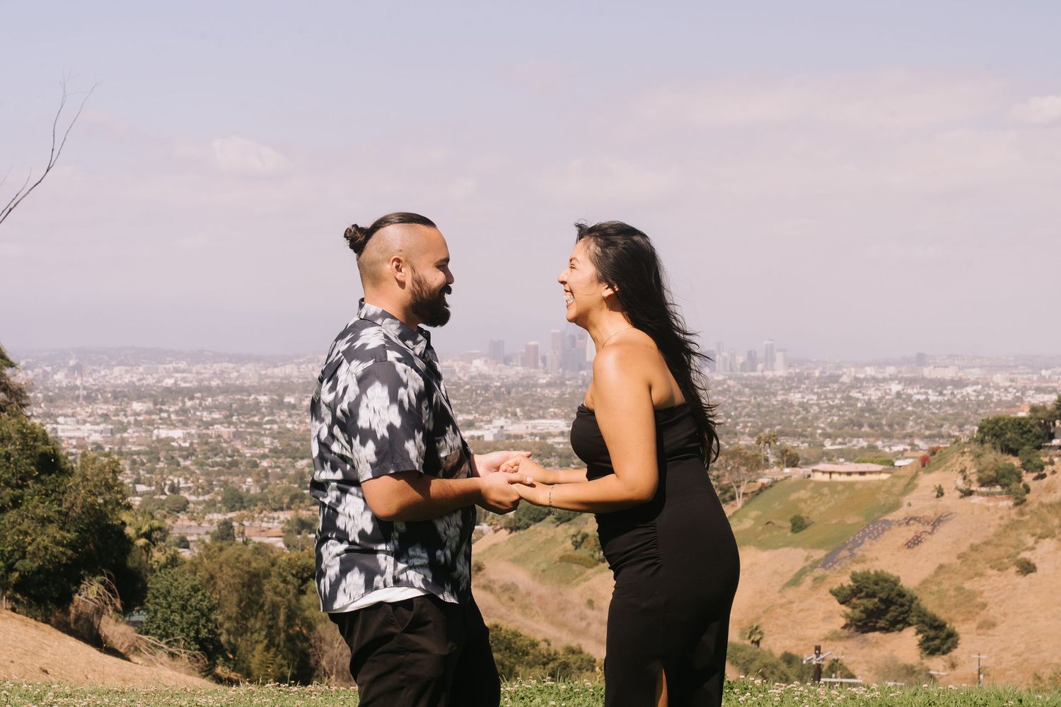 Couple holding hands and laughing on a hilltop with Los Angeles skyline in the background.