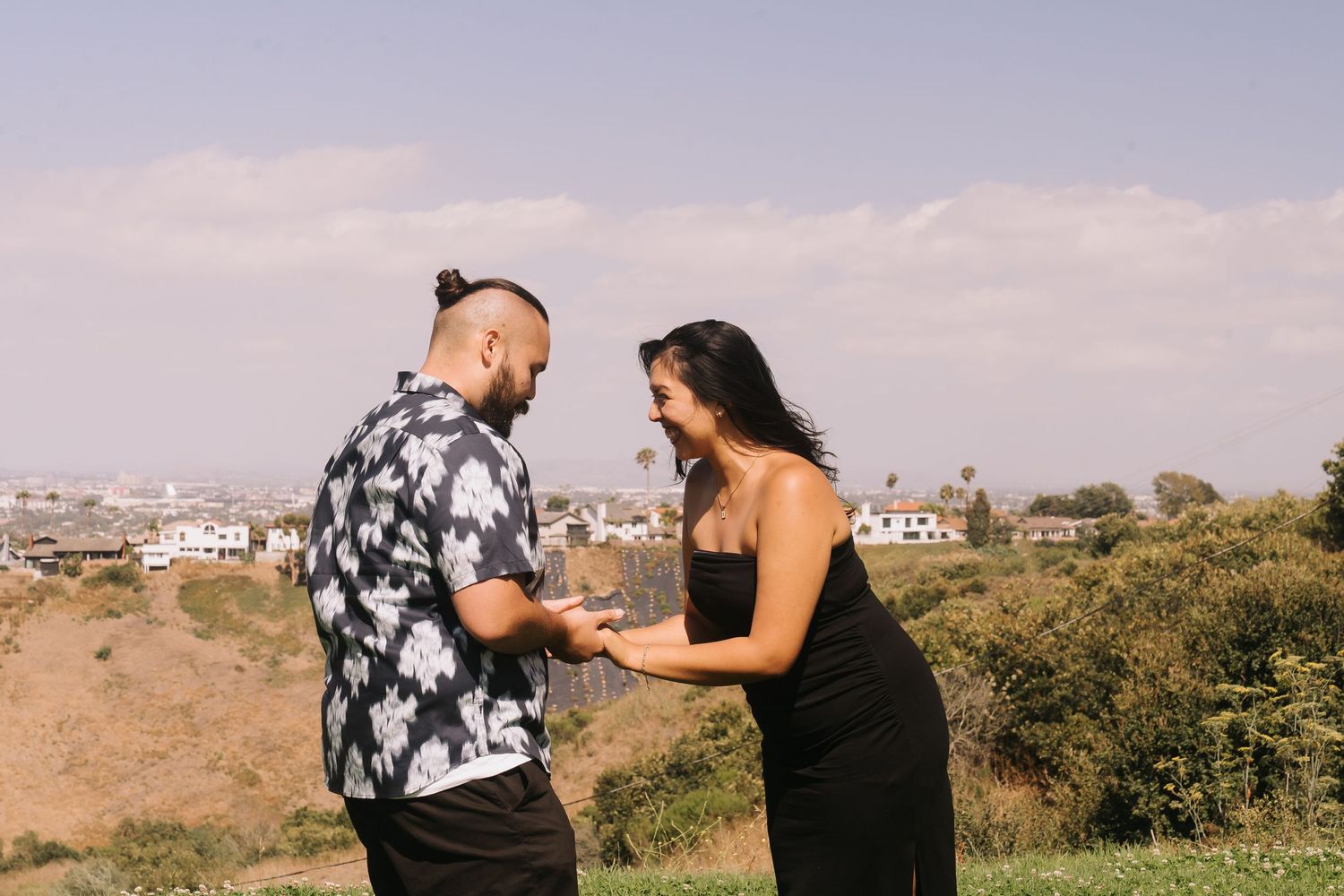 Couple sharing a romantic moment outdoors on a hilltop with a scenic cityscape in the background.