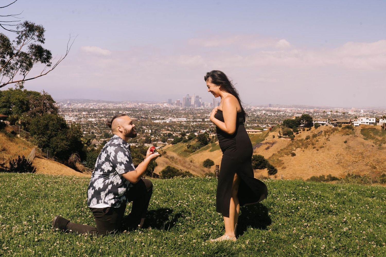 Man proposes on one knee to woman in black dress on grassy hill with Los Angeles skyline backdrop.