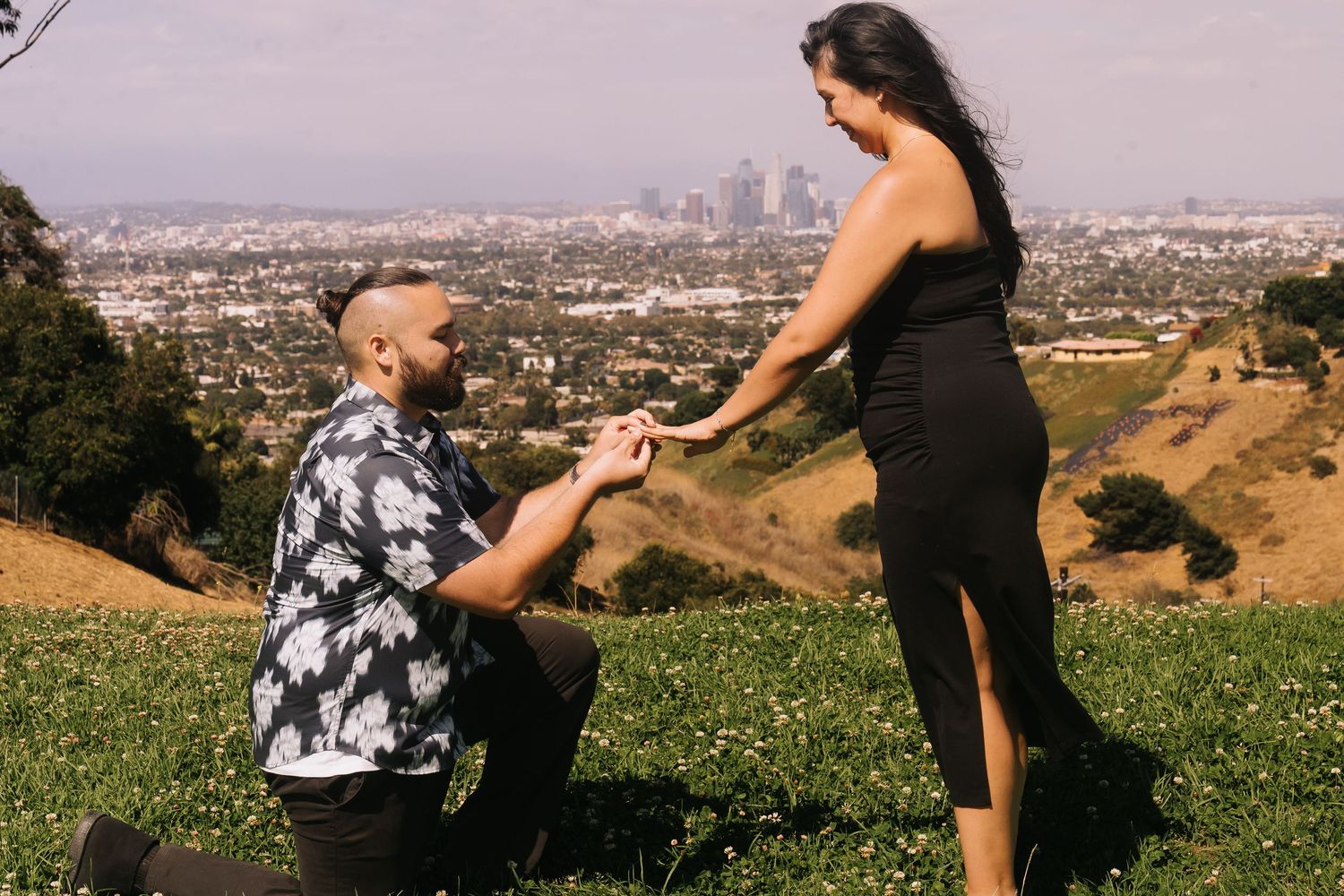 Man proposes on one knee to woman in black dress on a hillside with Los Angeles city skyline in background.