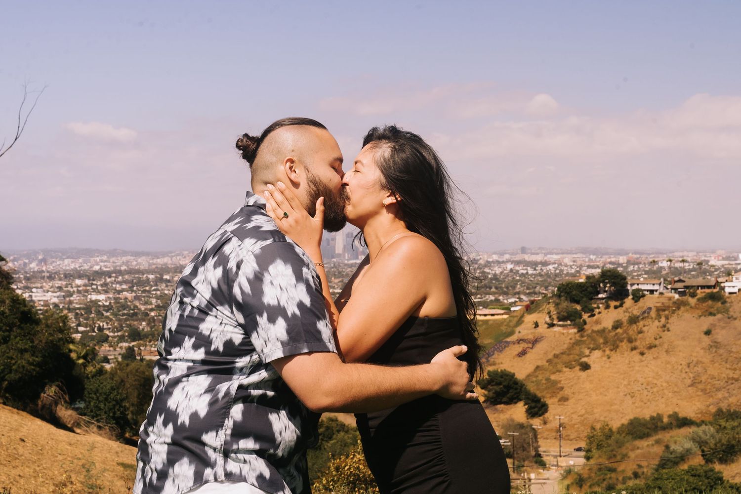 Couple sharing a romantic kiss on a hilltop overlooking a city skyline under a blue sky.
