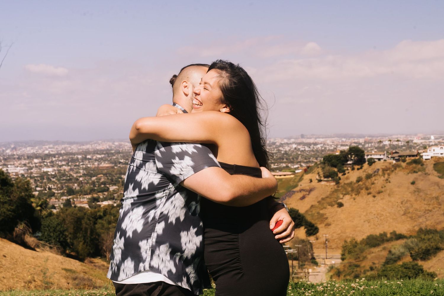 Two people share a warm embrace on a sunny hillside overlooking a sprawling city skyline.