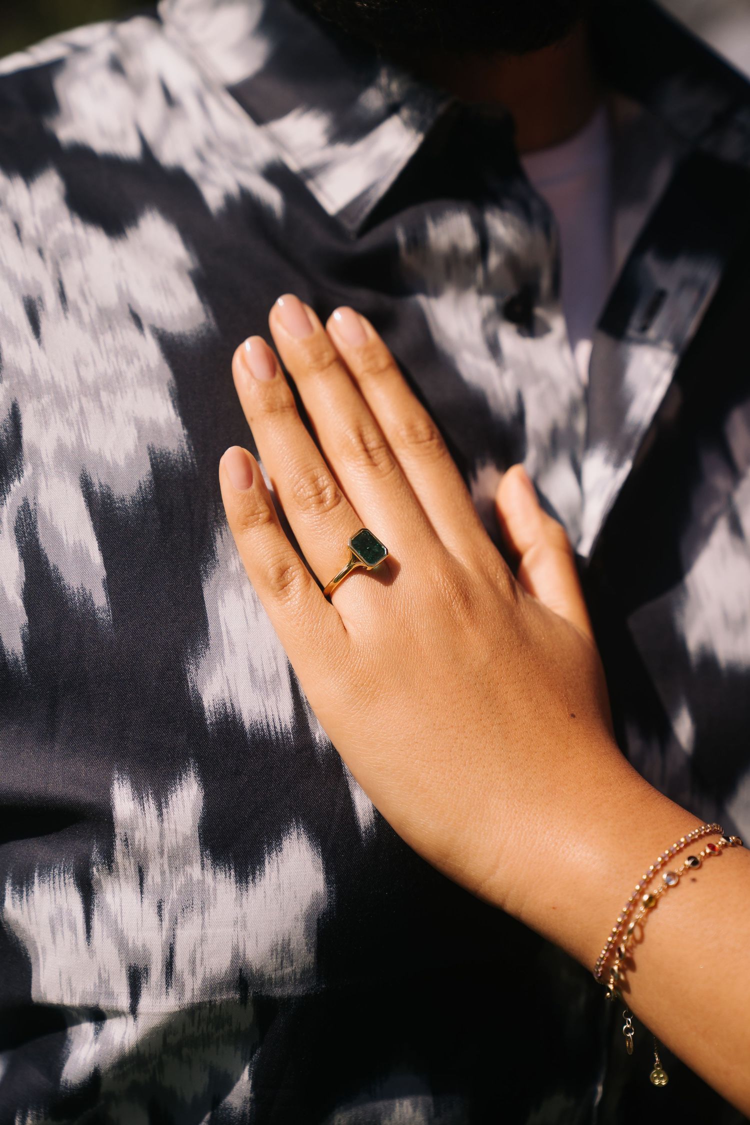 Close-up of a hand wearing a gold ring with a green stone and a delicate gold bracelet on a floral shirt.