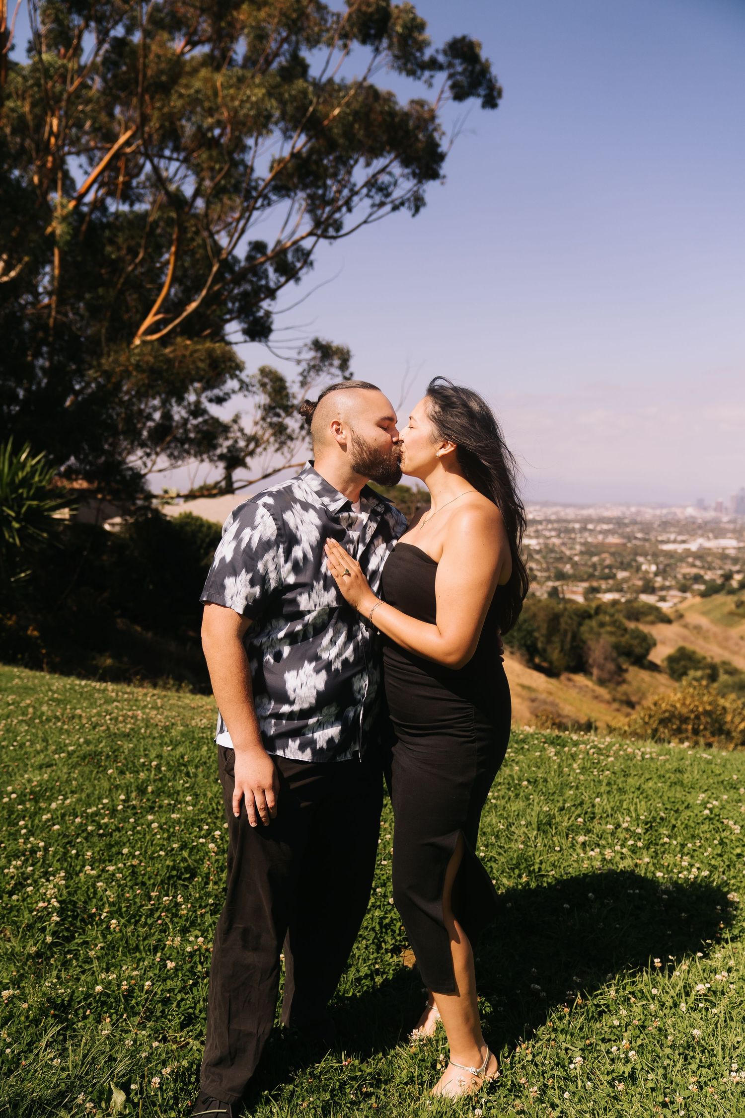Couple sharing a romantic kiss on a sunny hilltop with a panoramic city view and lush greenery behind them.