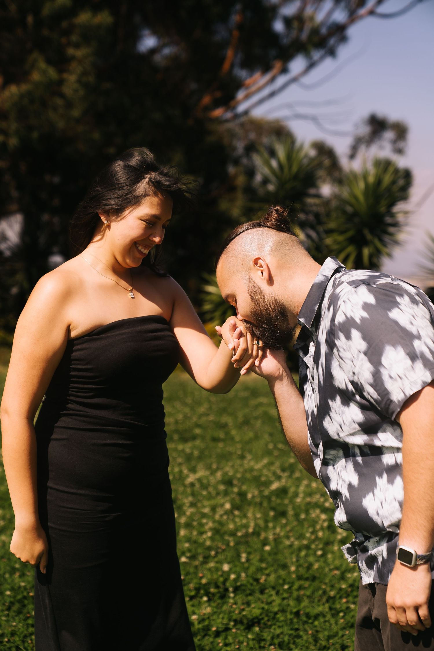 A man in a floral shirt kisses a woman's hand in a sunny park, as she smiles in a black strapless dress.
