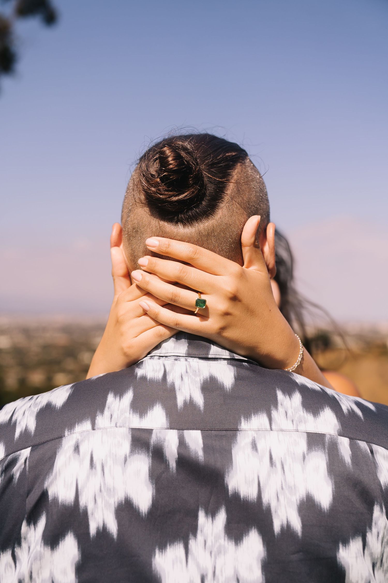 Back view of man with hair bun and undercut, woman's hand with green gemstone ring resting on his neck.