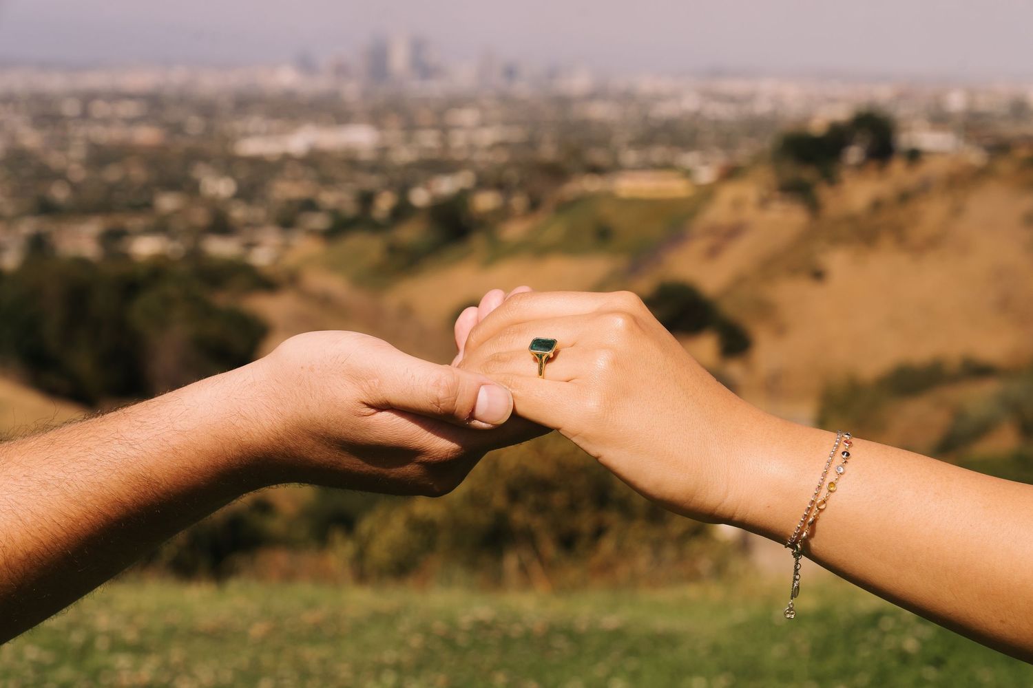 Two hands gently touching with a gold ring and bracelet, hills and city skyline in the soft background.
