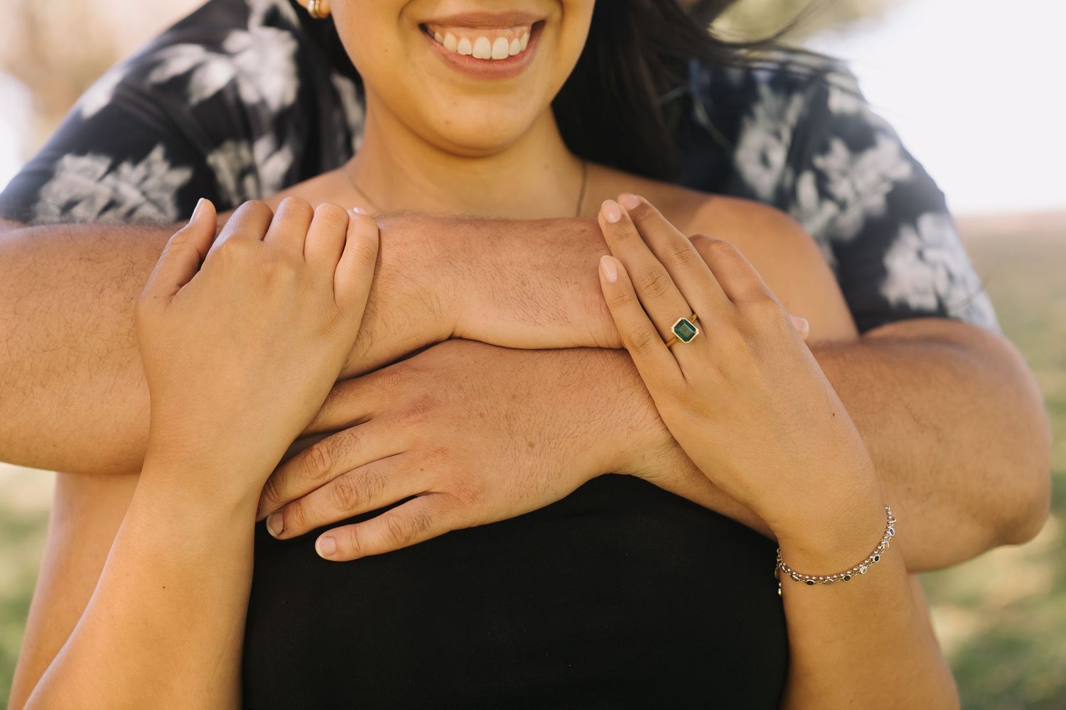 Smiling woman in black top with a man's arms wrapped around her, showcasing a green gemstone ring and gold bracelet.