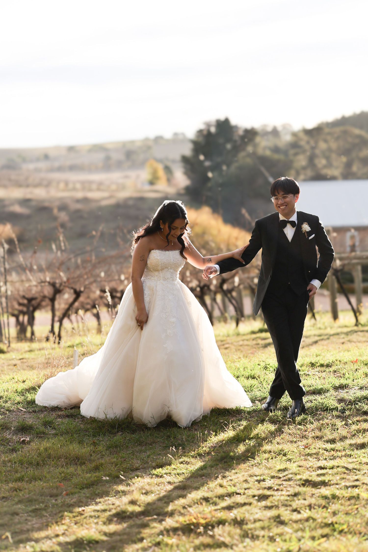 Couple in wedding attire walking hand in hand through a vineyard, bride in ball gown and groom in black tuxedo.