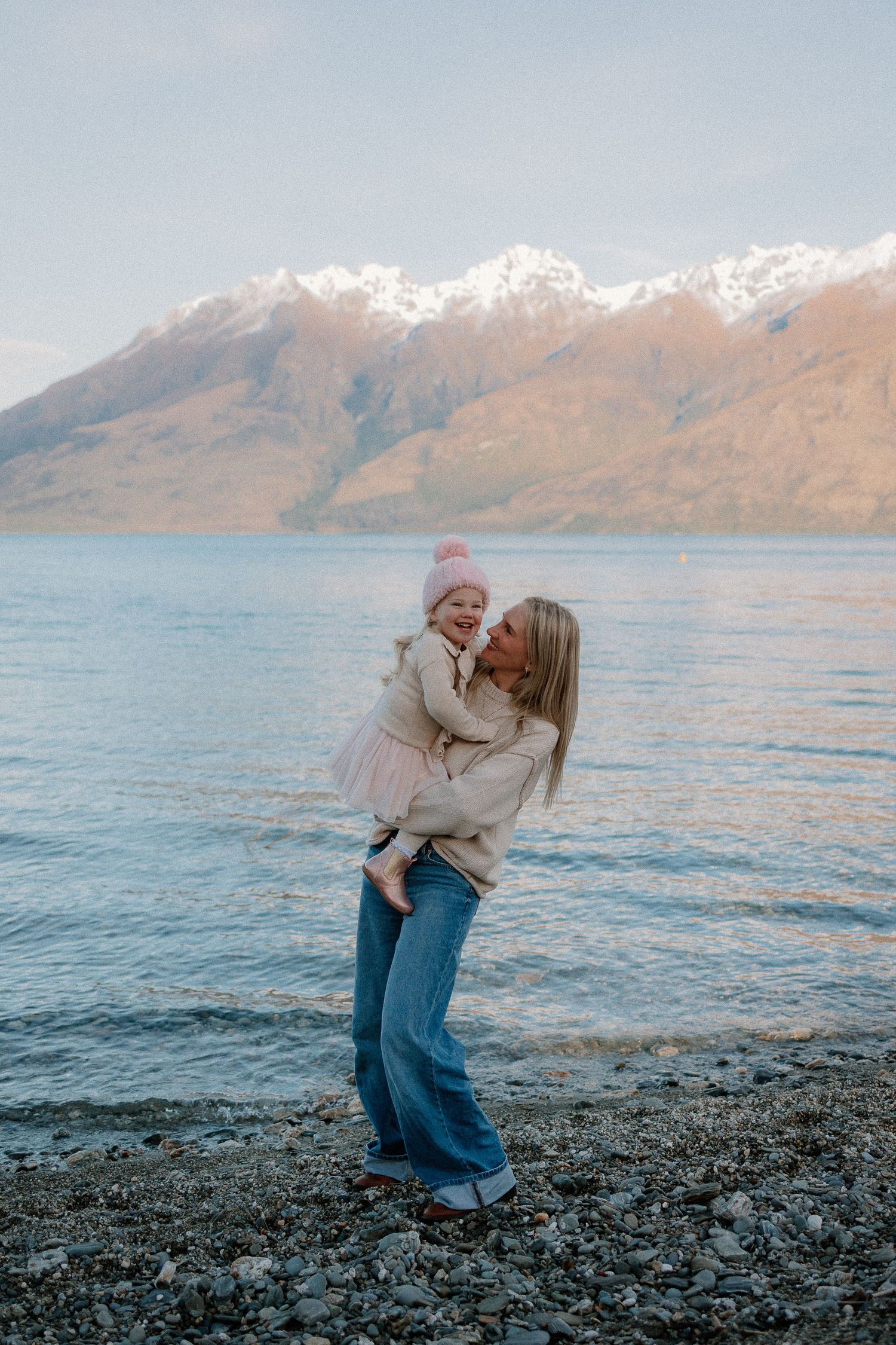 A woman holds a baby by a rocky lakeside with snow-capped mountains in the background.