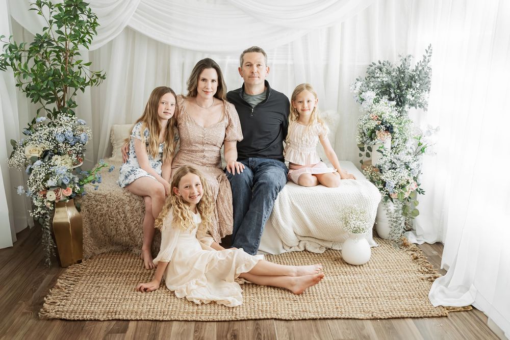 Family of five posing for a portrait in a bright floral studio setting with white drapes and greenery.