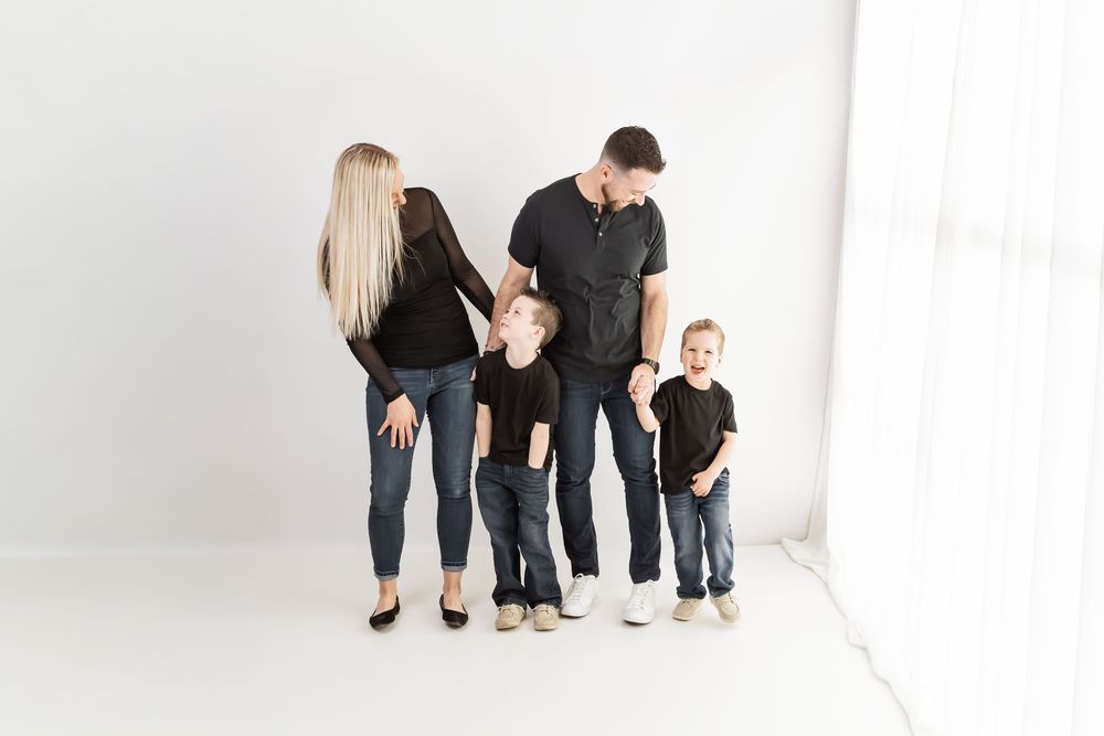 Family of four in matching black outfits posing together in a bright white studio, parents with two young boys.