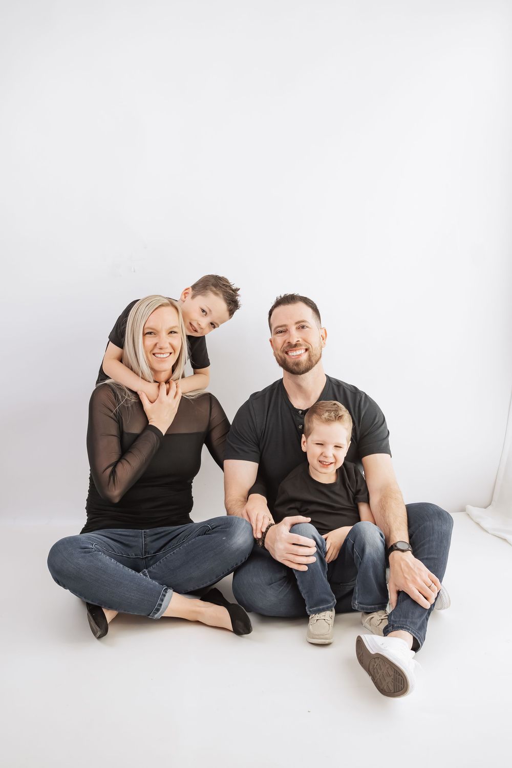 Happy family of four in matching black tops and jeans sitting together on white background, smiling at camera.