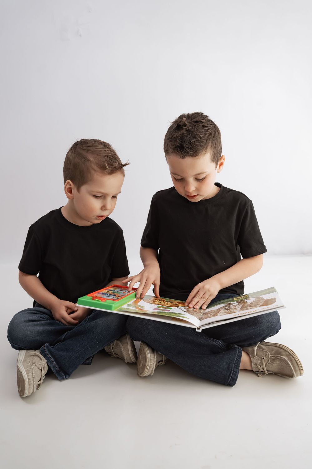 Two young boys in black t-shirts sitting cross-legged on floor reading a colorful picture book together.