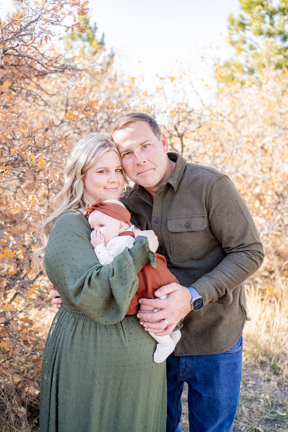 A couple smiles holding their newborn baby outdoors among autumn foliage, wearing green and rust tones.