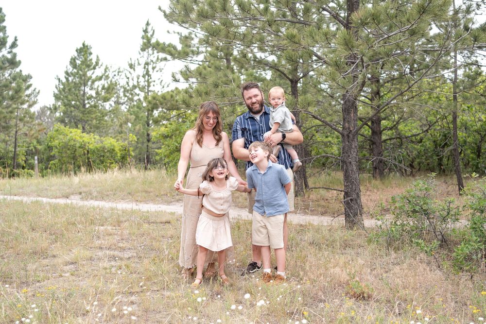 Family of five posing outdoors in a pine forest meadow, parents with three young children smiling together.