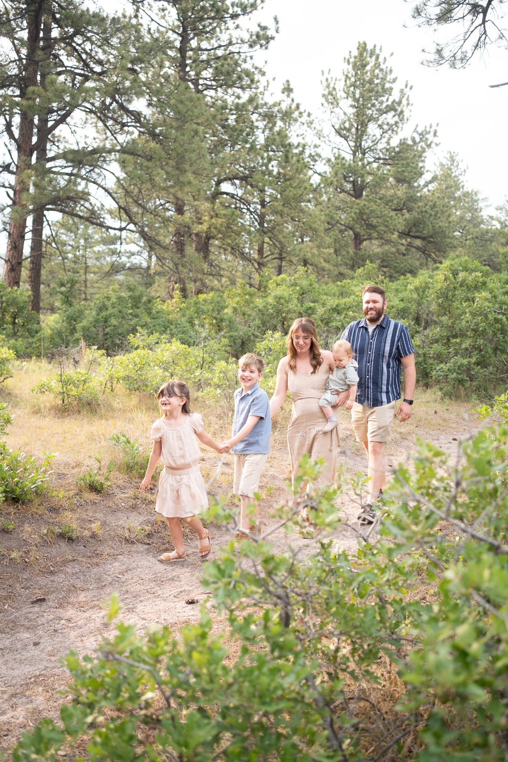 Happy family of five walking together on a forest trail surrounded by pine trees and lush greenery.