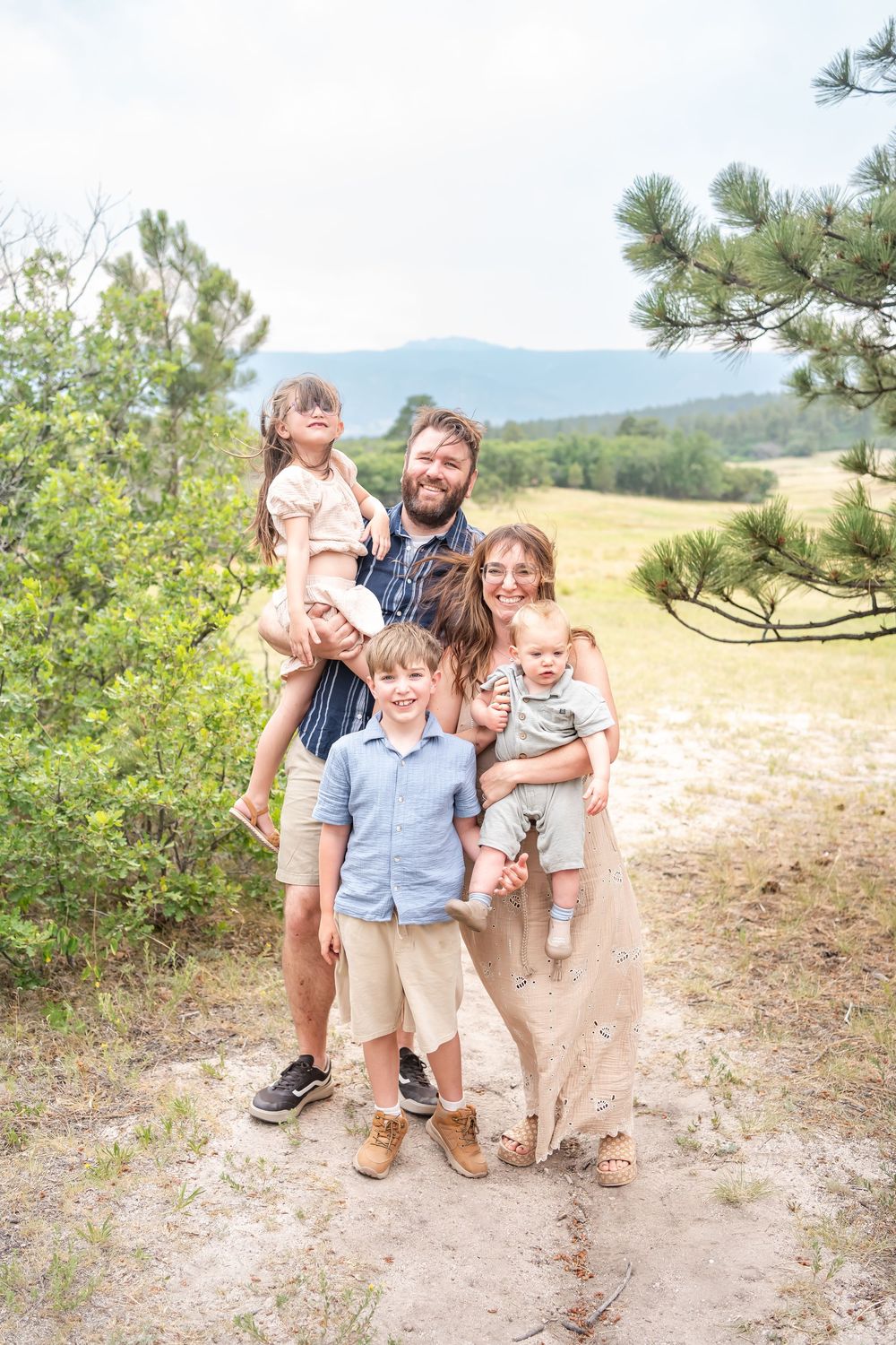 Happy family of six posing outdoors on a sandy trail surrounded by pine trees and mountain meadows.