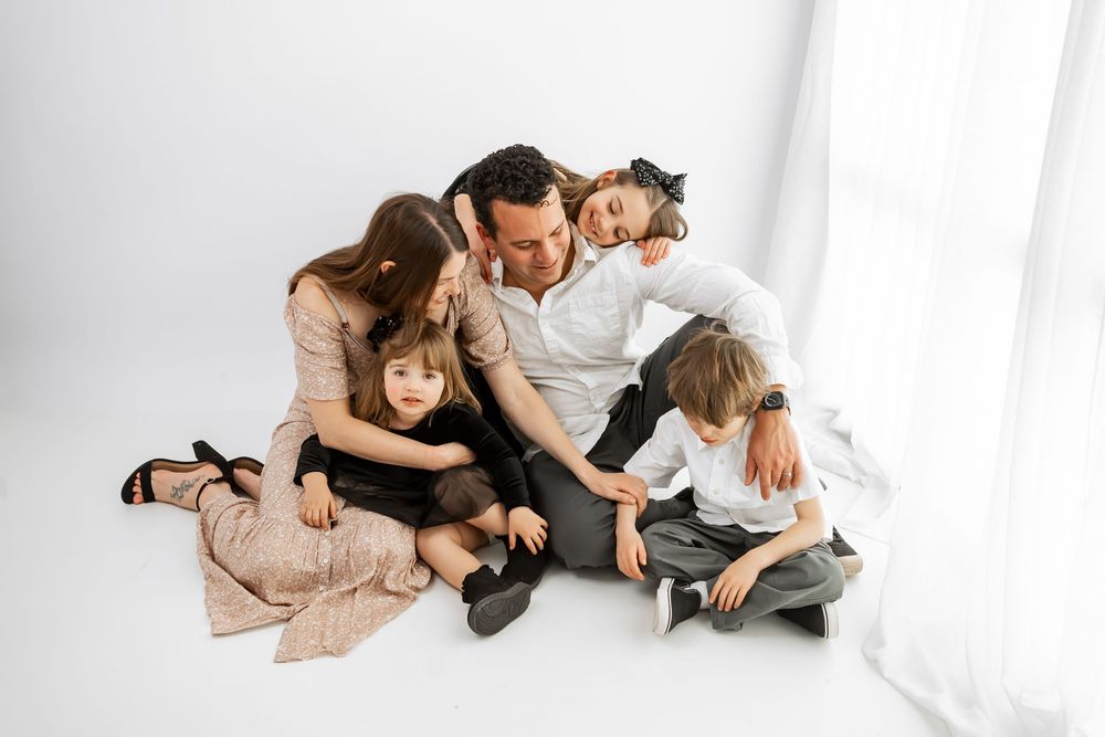 A happy family of five sits together on a white studio floor, sharing warm embraces and smiles.