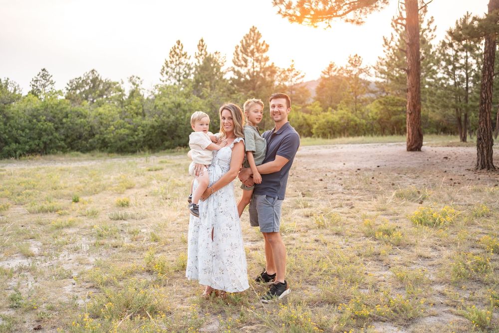 Happy family of three posing outdoors at golden hour in a sunny meadow surrounded by pine trees.