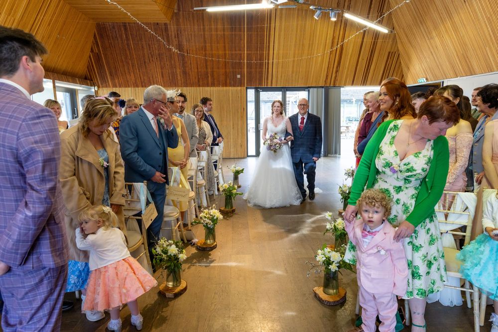 Bride walks down the aisle with escort in a wooden chapel, guests standing and smiling, flower arrangements lining the path.