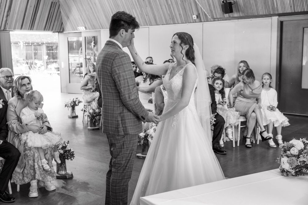 Bride and groom exchange vows at indoor wedding ceremony with seated guests in black and white photo.
