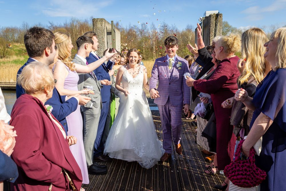 Bride and groom in purple suit exit dock amid confetti shower with cheering wedding guests.