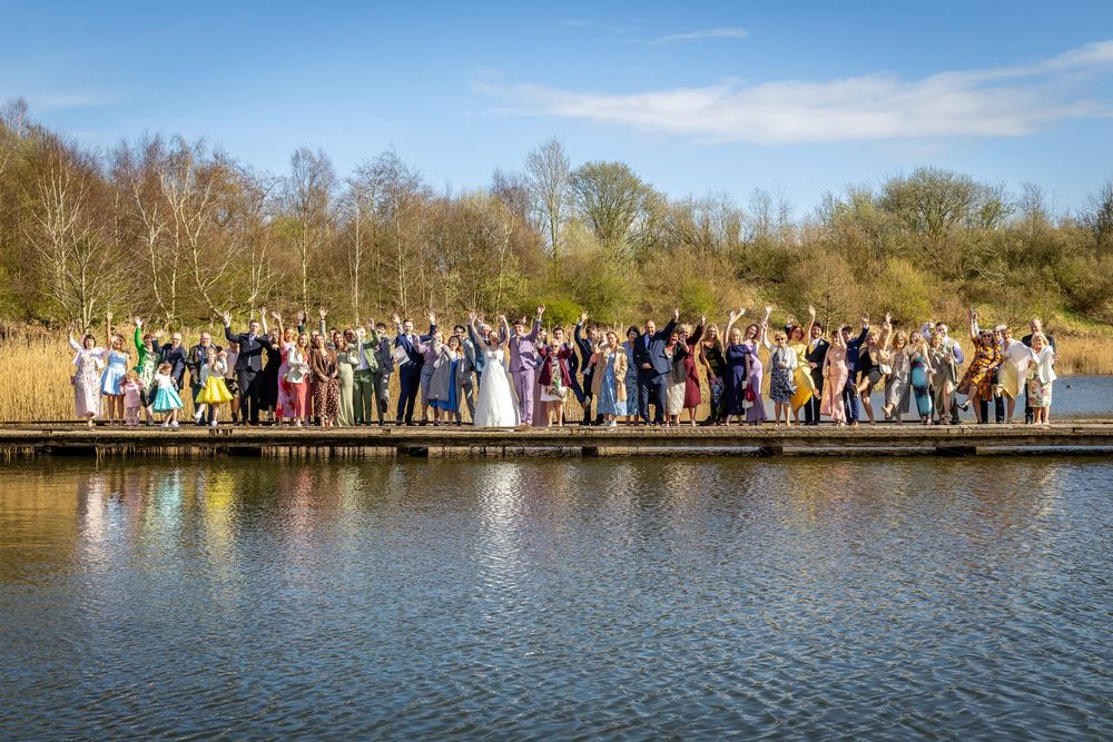 Large wedding party celebrating on a wooden dock by a serene lake surrounded by lush trees under a bright blue sky.