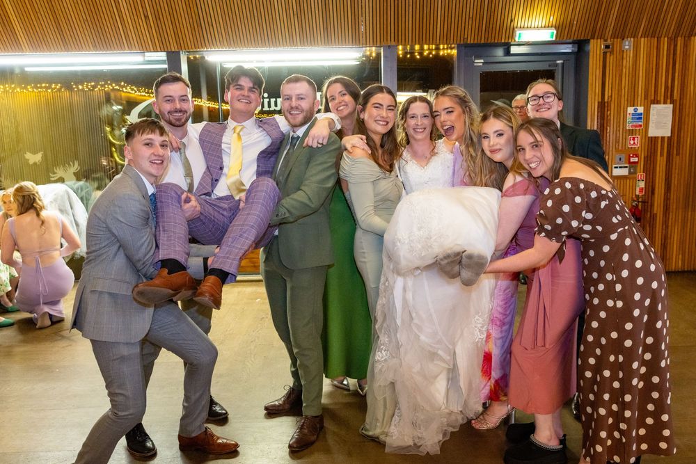 Wedding party celebrating joyfully, bride in white gown lifted by friends on dance floor at festive indoor reception.