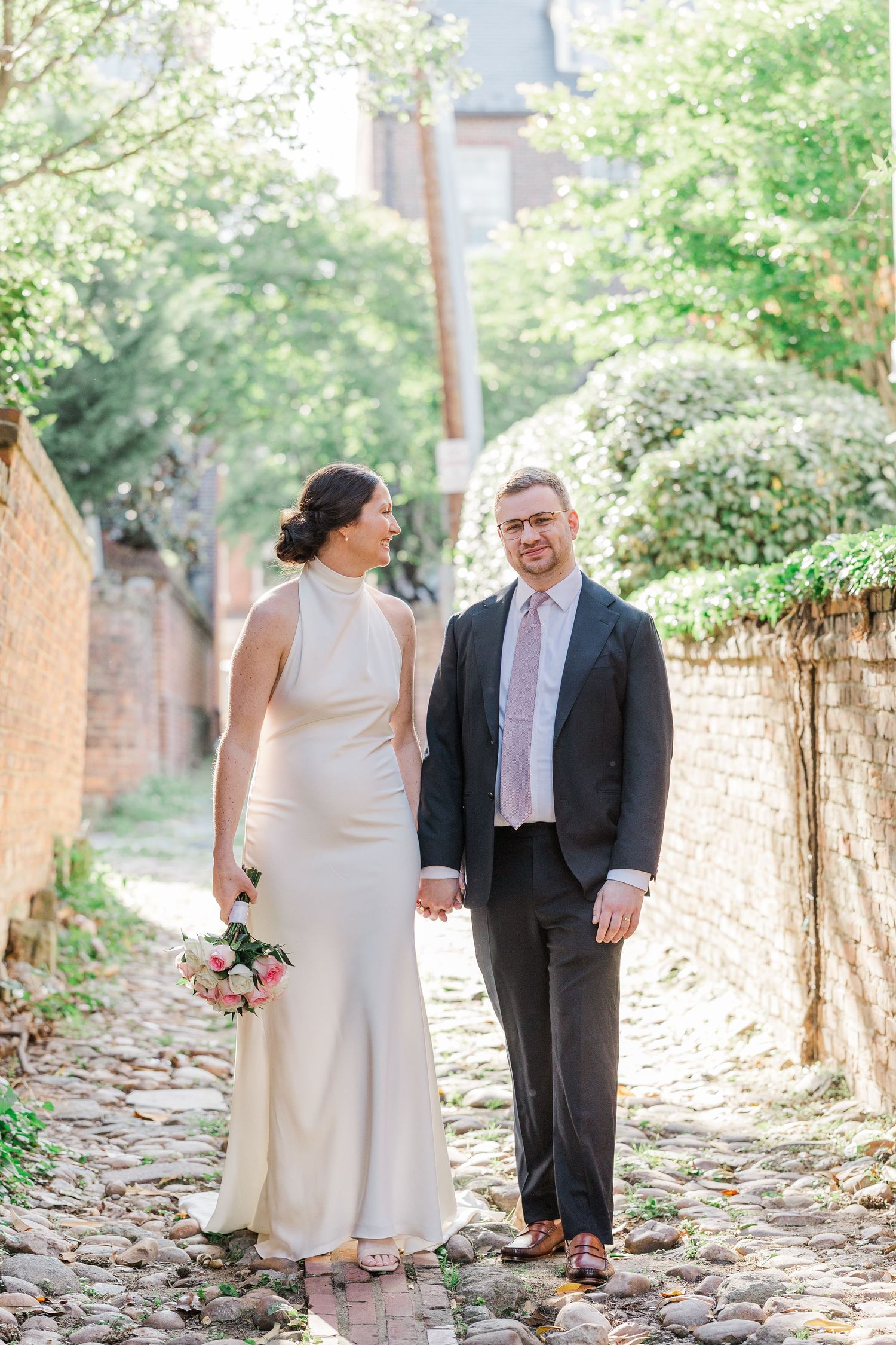 Bride and groom holding hands walking on cobblestone path, bride holding floral bouquet.