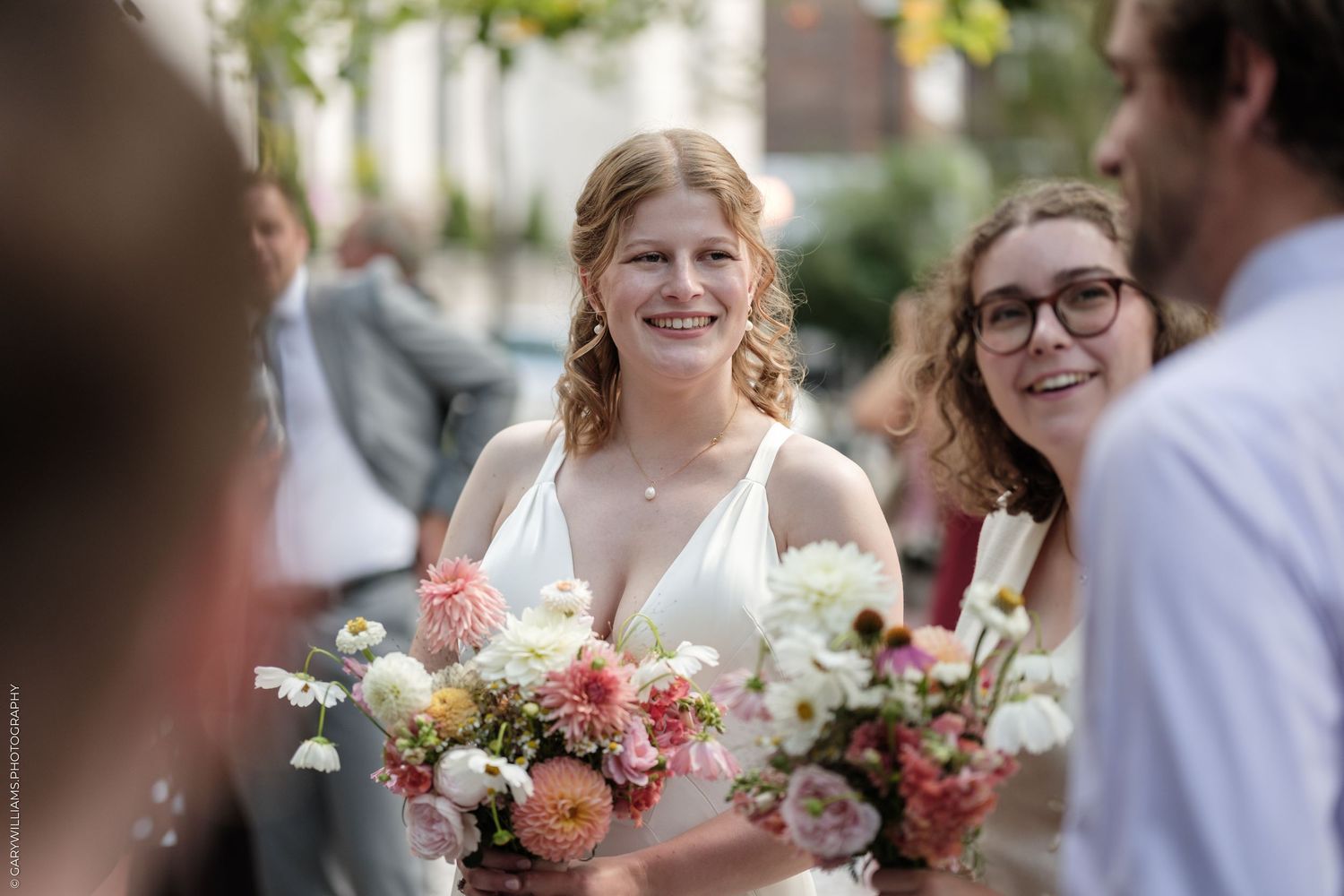 Smiling bride in white dress holding colorful wedding bouquet surrounded by guests outdoors on her wedding day.