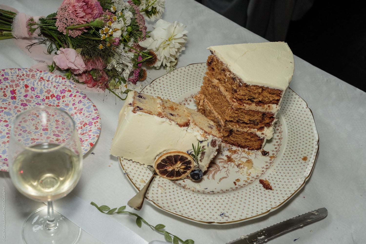 Slice of layered carrot cake with white frosting on floral plate, wine glass and pink flowers in background.