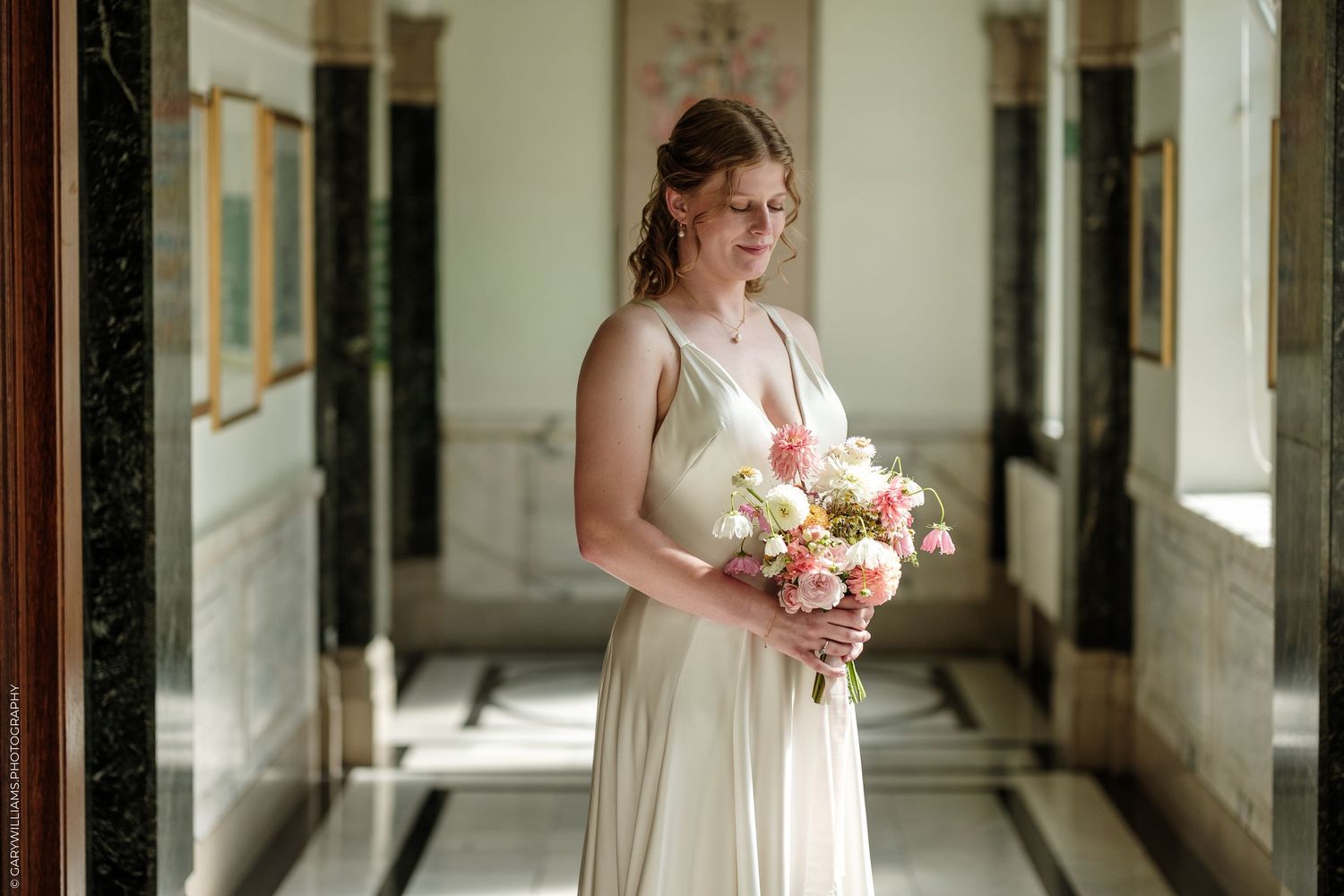 Bride in ivory gown holds pink and white bouquet in elegant marble hallway.