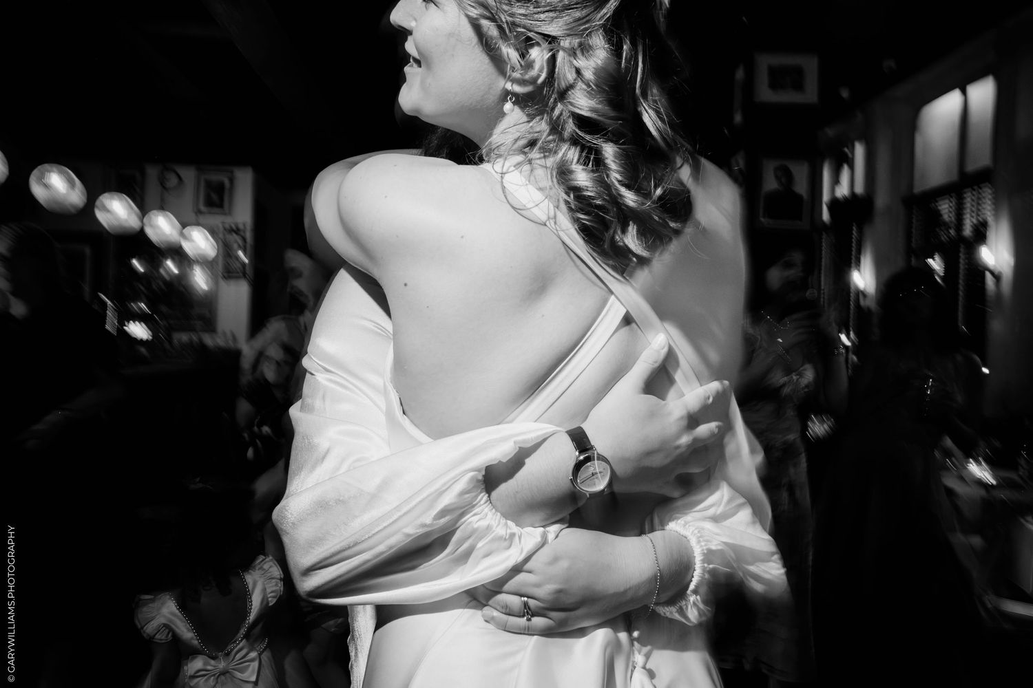 Black and white photo of a bride in a backless wedding gown being embraced at a reception.