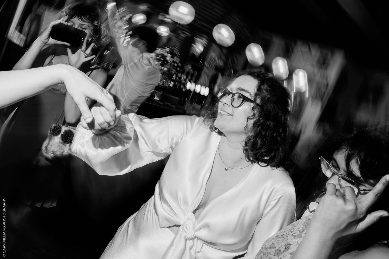 Black and white photo of a smiling woman with curly hair and glasses at a lively social gathering.